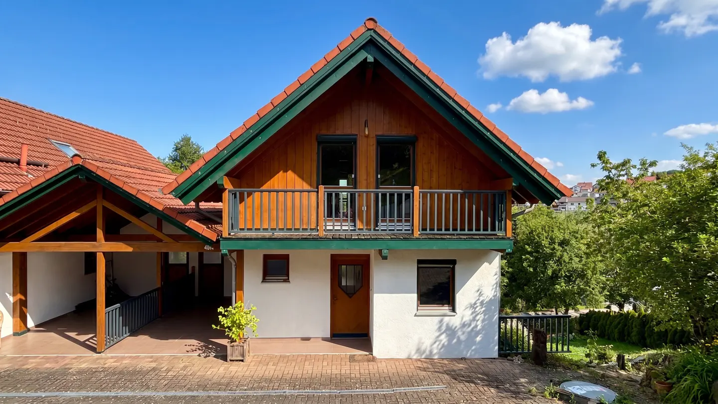 Two-story house with a red tile roof, green trim, and a wood balcony on the second floor. The house has a white exterior and a brown front door.