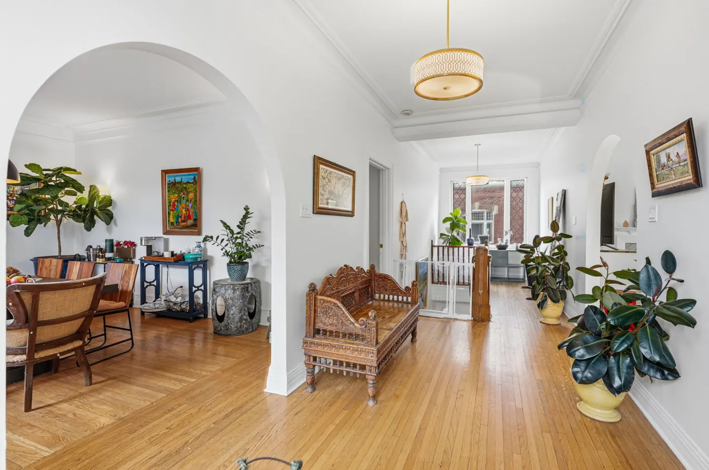 A bright hallway with hardwood floors, white walls, and arched doorways. A carved wooden bench sits in the center, with plants and artwork throughout.