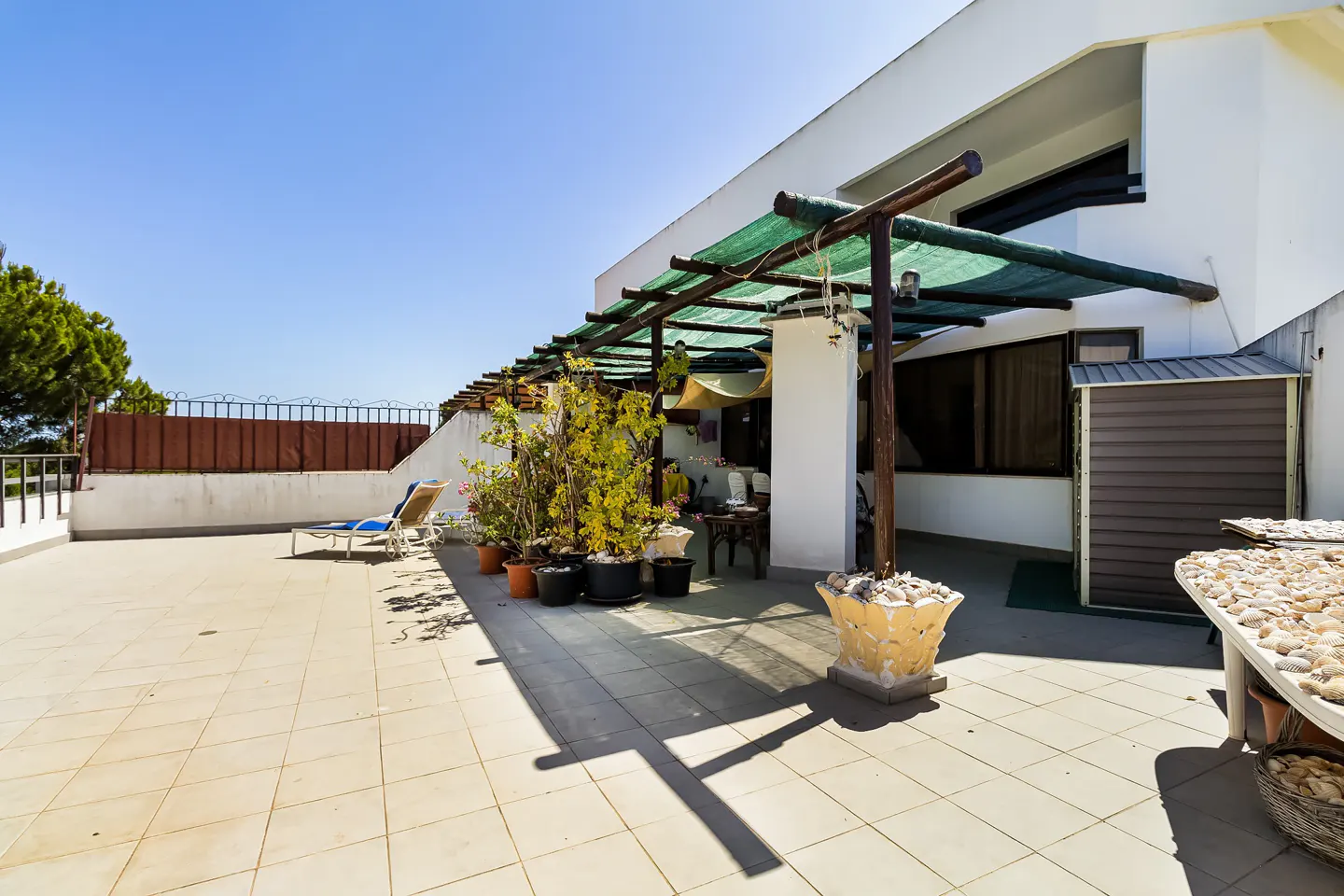 A sun-drenched patio with tile flooring, a pergola with green shade, potted plants, and a lounge chair.