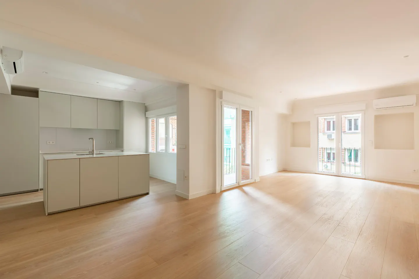Bright, empty apartment with light wood floors. A gray kitchen island sits to the left, and windows overlook a city street.