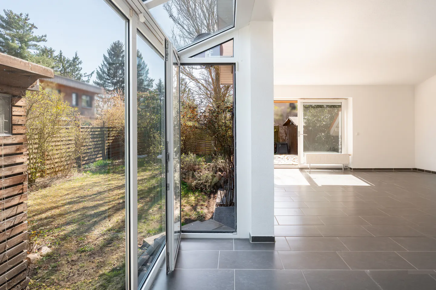 Bright, empty room with gray tile floor and white walls. Open glass doors lead to a green yard with trees and a wooden fence.