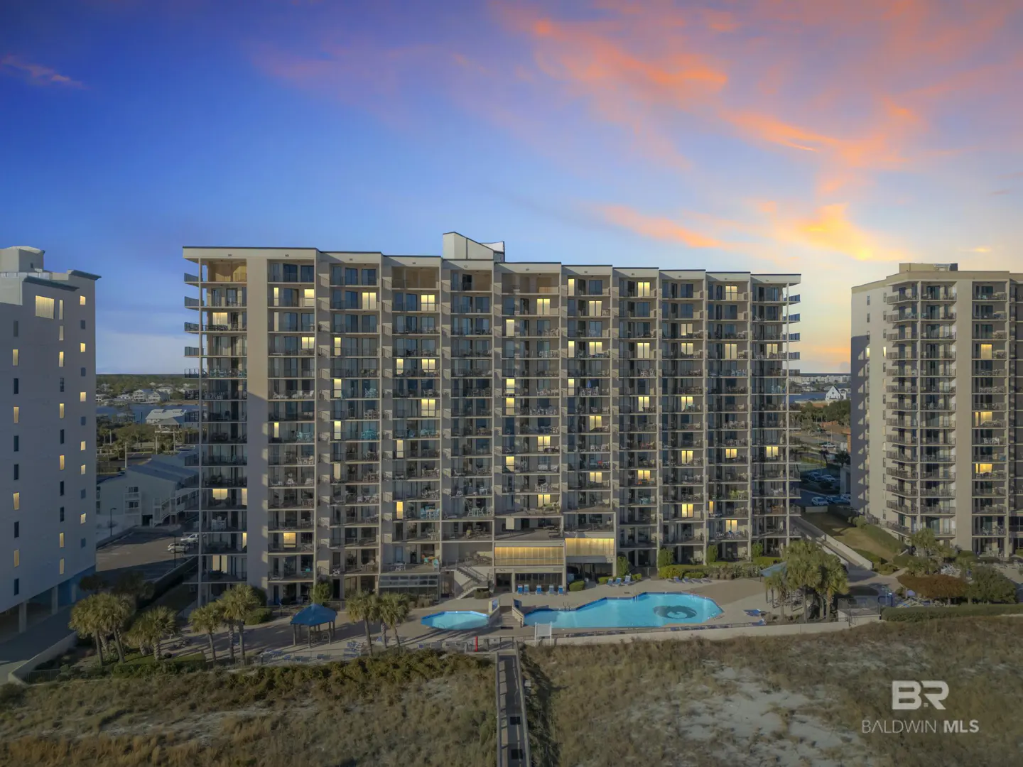 Exterior view of a tall condo building with a pool, palm trees, and beach access under a colorful sunset sky.