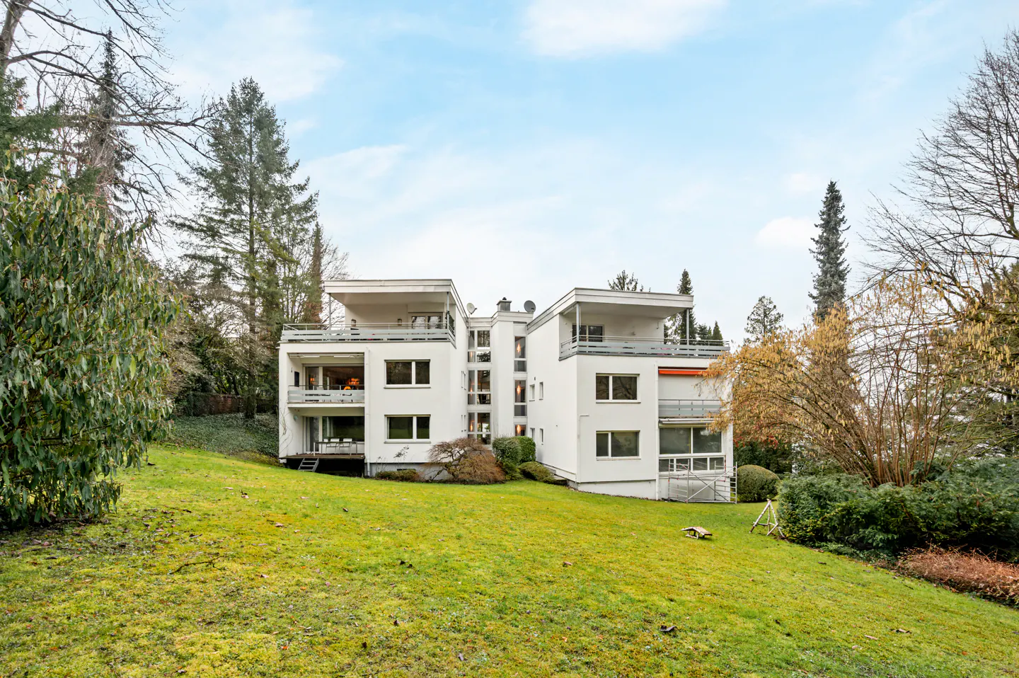Modern white apartment building with balconies on a green lawn, surrounded by trees under a blue sky.