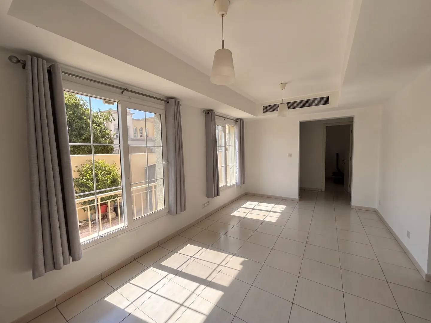 Bright, empty room with white walls, tile floor, and two windows with gray curtains. Light fixtures hang from the ceiling.