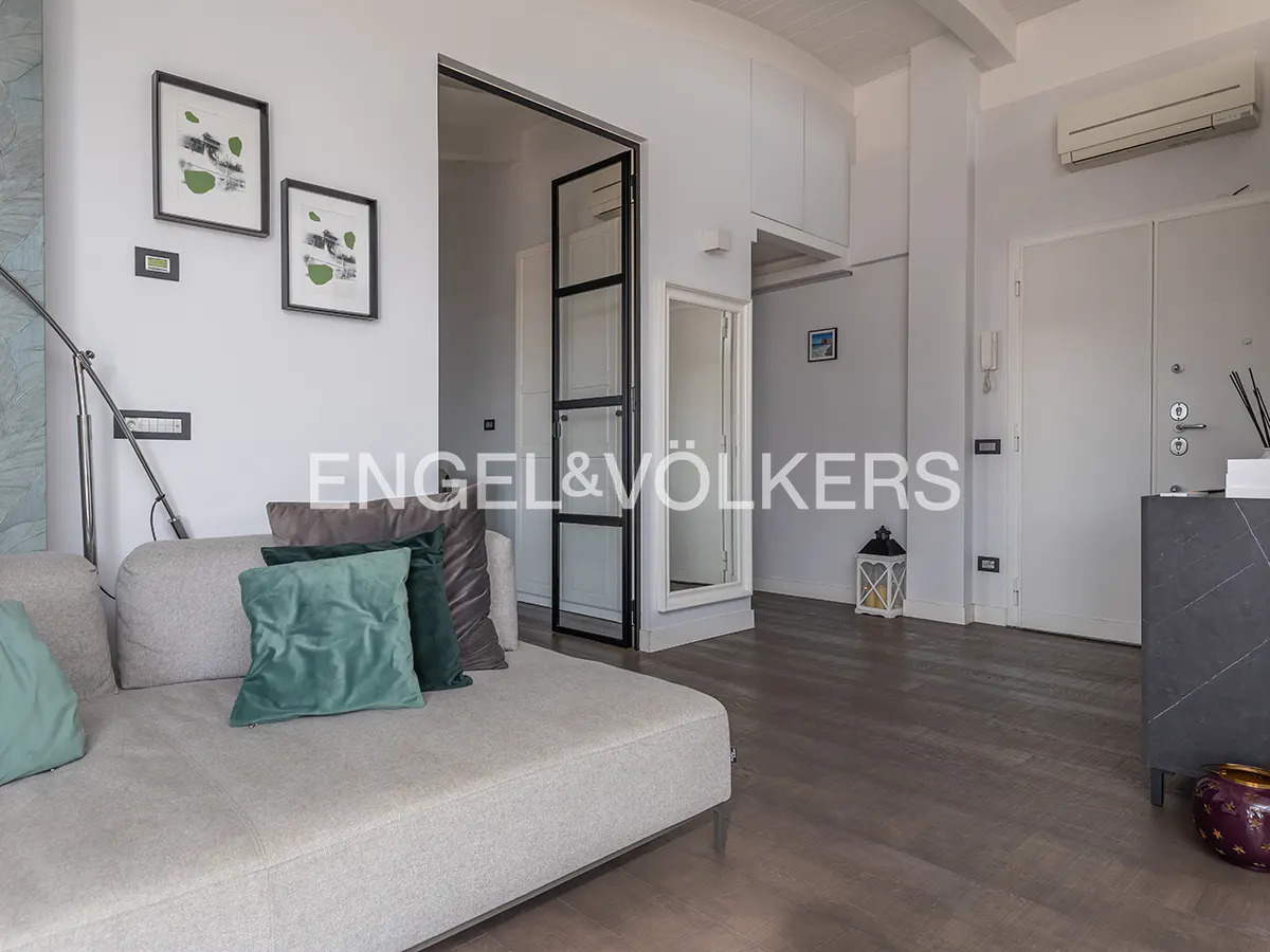 Living room with a gray sofa, green pillows, and artwork on the white walls. A black-framed glass door leads to another room.