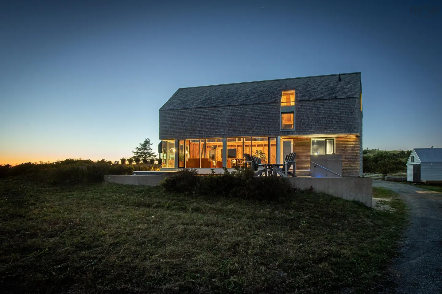 Modern two-story home with gray shingles and large windows at dusk. Lawn in foreground.