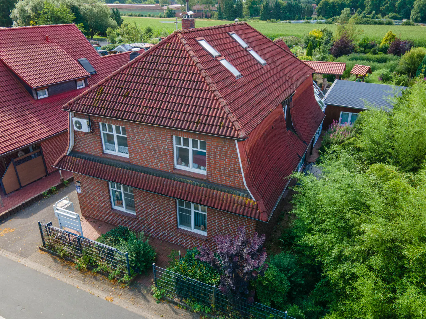 Aerial view of a two-story brick house with a red tile roof and white-framed windows, surrounded by green trees and bushes.