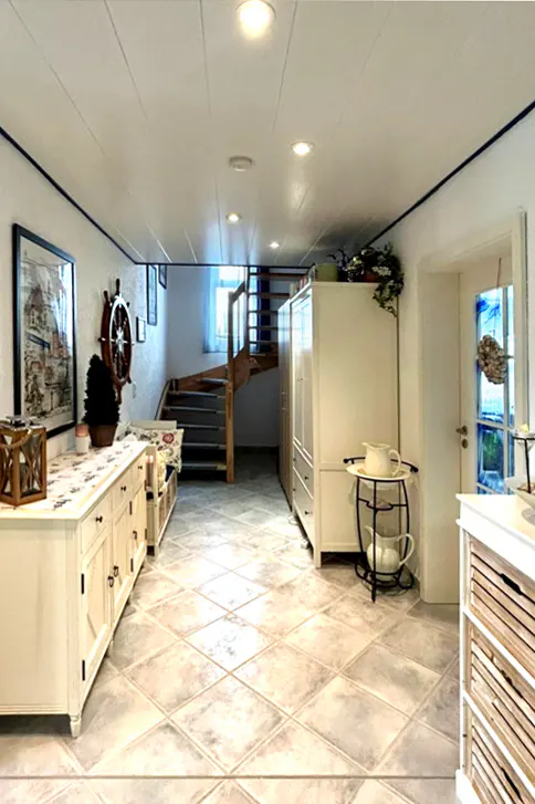 Bright hallway with tile floors, white cabinets, and a wooden staircase. A ship's wheel hangs on the wall. Natural light streams through a door.