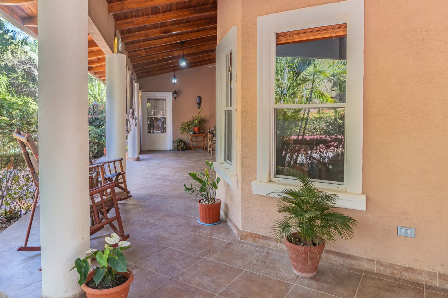Covered porch with rocking chairs, white columns, and potted plants. A window reflects lush greenery. The ceiling is wood.