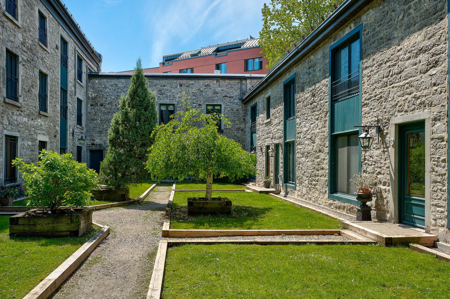 Courtyard view of stone buildings with green trim, grass, trees, and gravel paths under a blue sky.