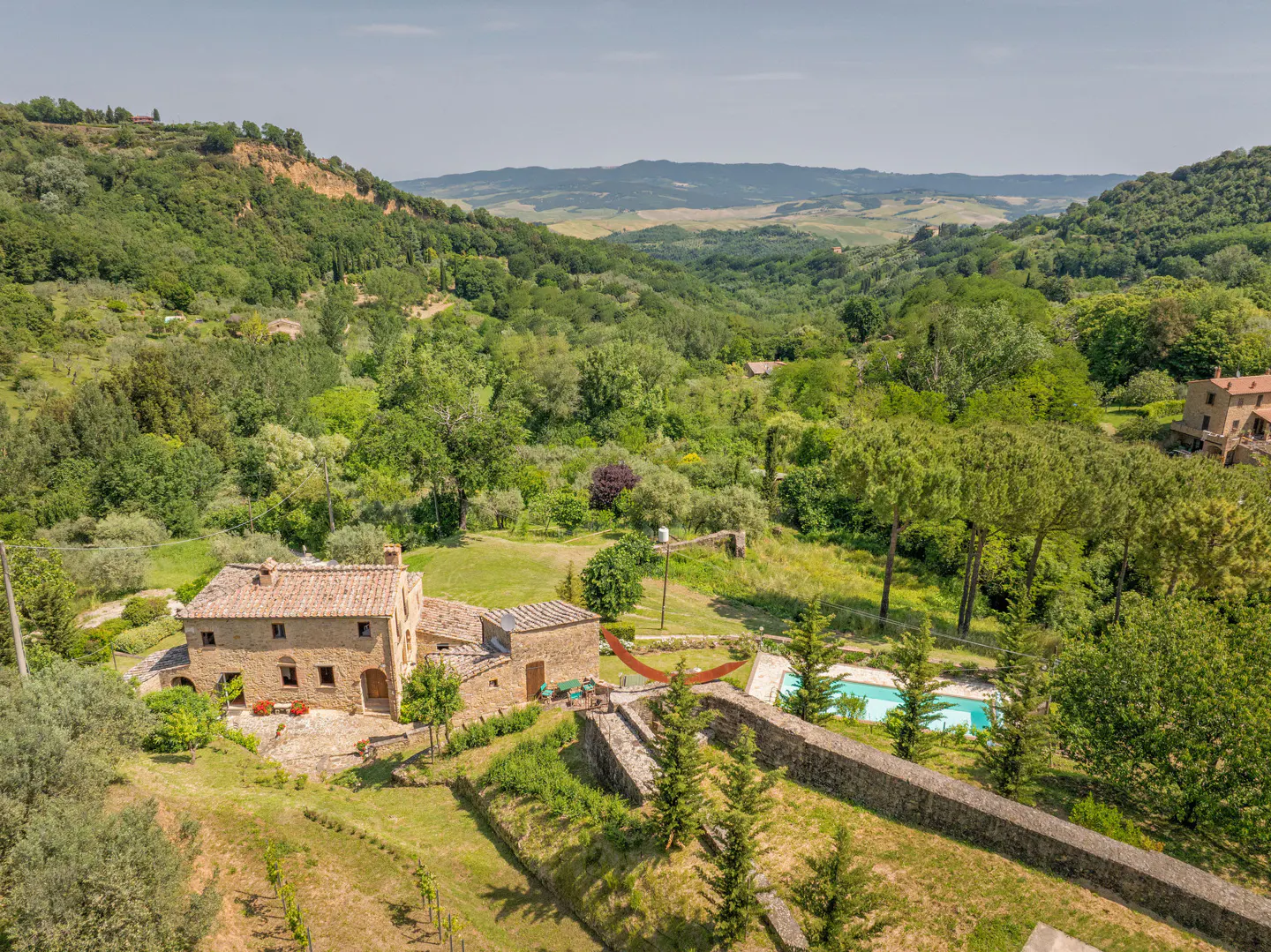 Aerial view of a stone villa in the Tuscan countryside with a swimming pool, surrounded by lush hills and trees.