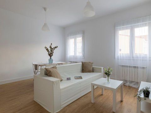 Bright, all-white living room with a sofa, table, and wood floors. Windows with sheer curtains let in natural light.