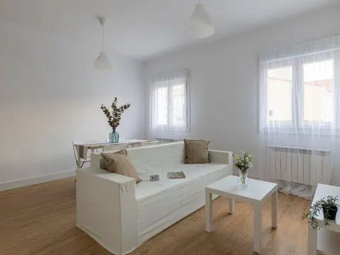 Bright, all-white living room with a sofa, table, and wood floors. Windows with sheer curtains let in natural light.