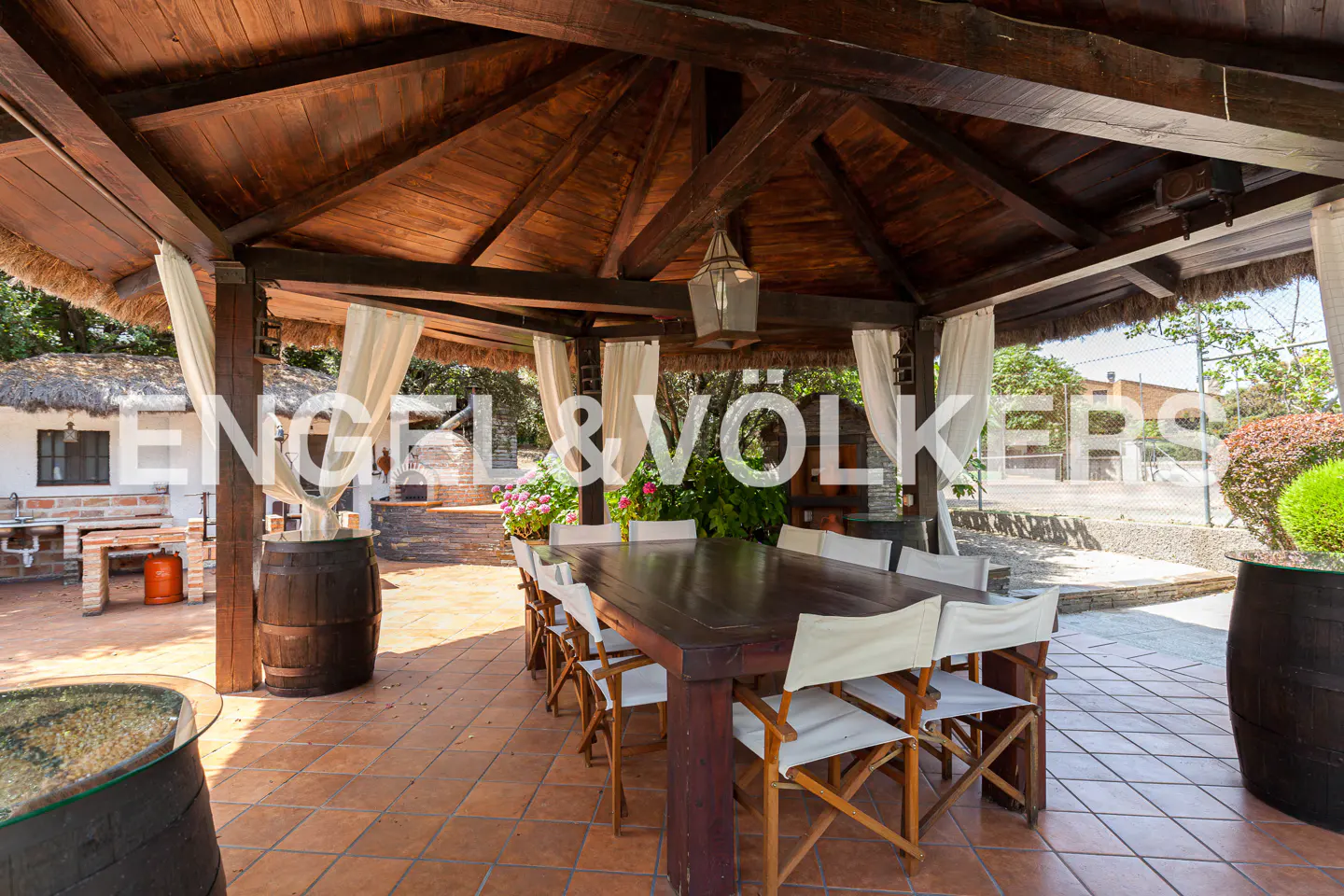 Outdoor dining area with a dark wood table and white chairs under a thatched roof gazebo with white curtains. Barrels and greenery surround the space.