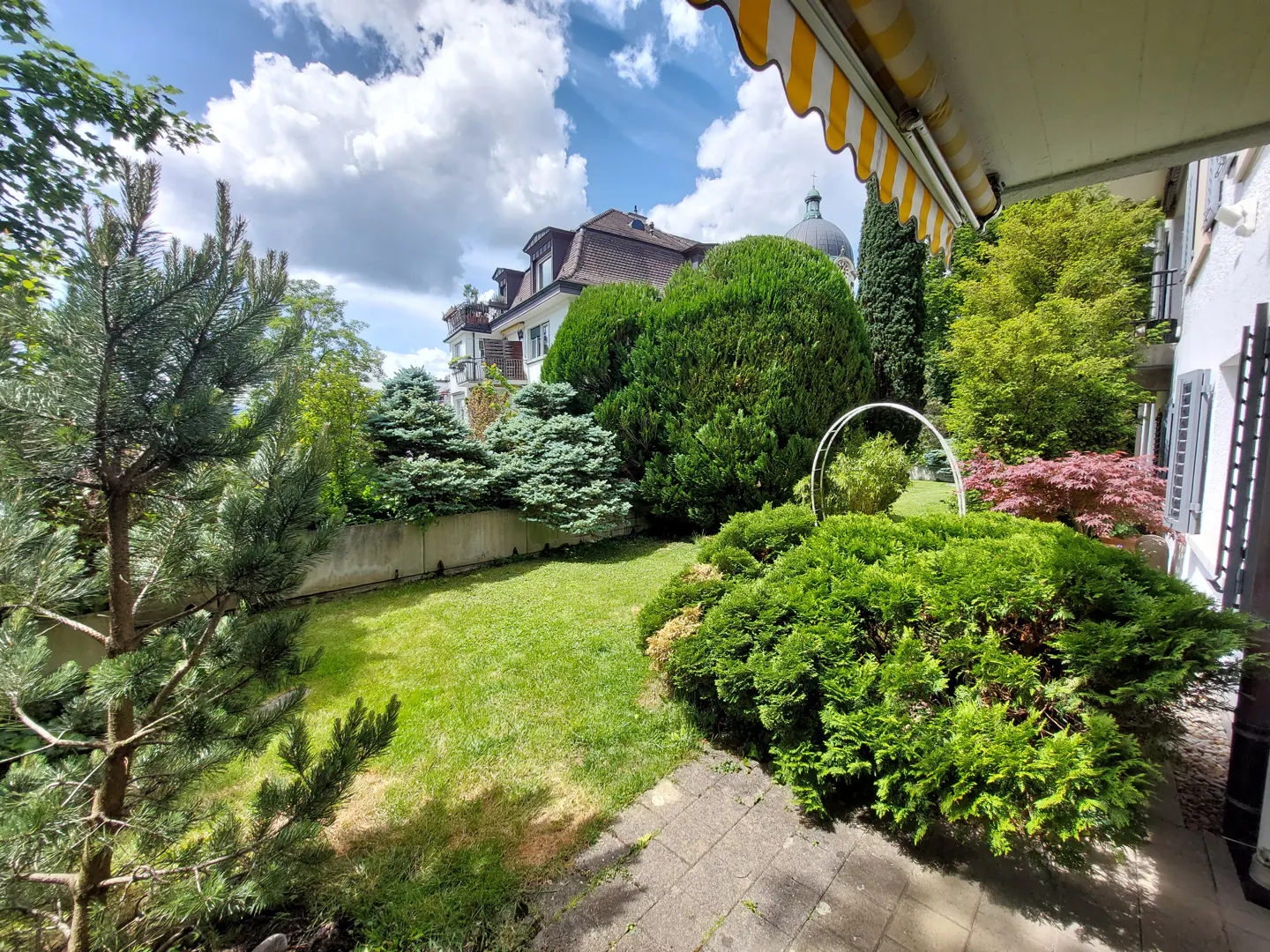 View of a lush green lawn and garden with trees, bushes, and a building in the background under a blue sky with white clouds.
