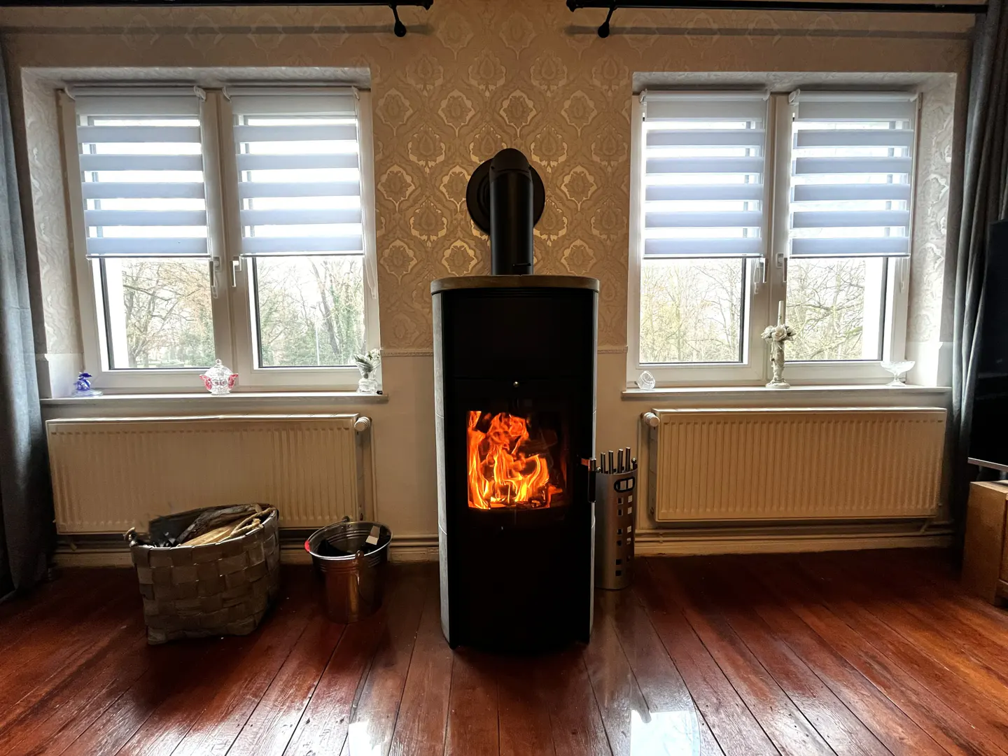 Living room with a black wood-burning stove with a fire burning inside, hardwood floors, and two windows with white blinds.