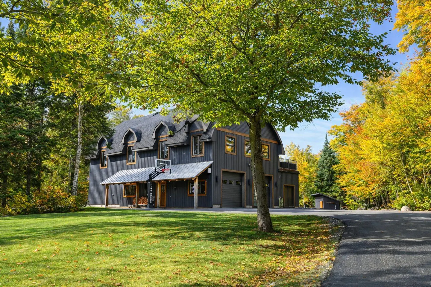 Exterior of a large, modern gray house with a basketball hoop, surrounded by green grass and fall foliage.