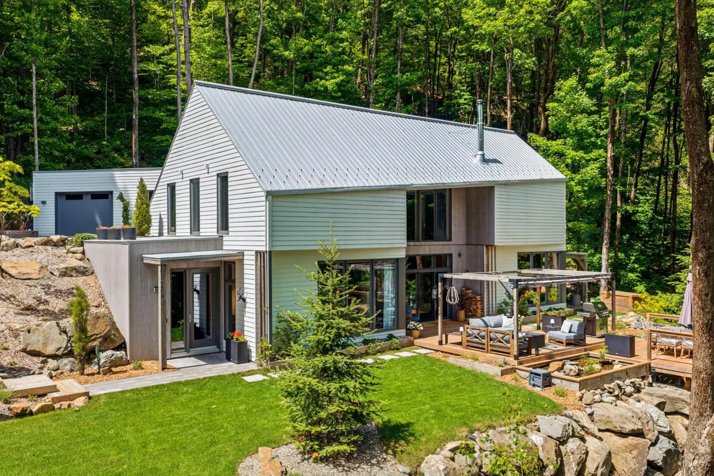 Modern white home with metal roof, wooden deck, and patio furniture, nestled in a lush green forest with rocky landscaping.