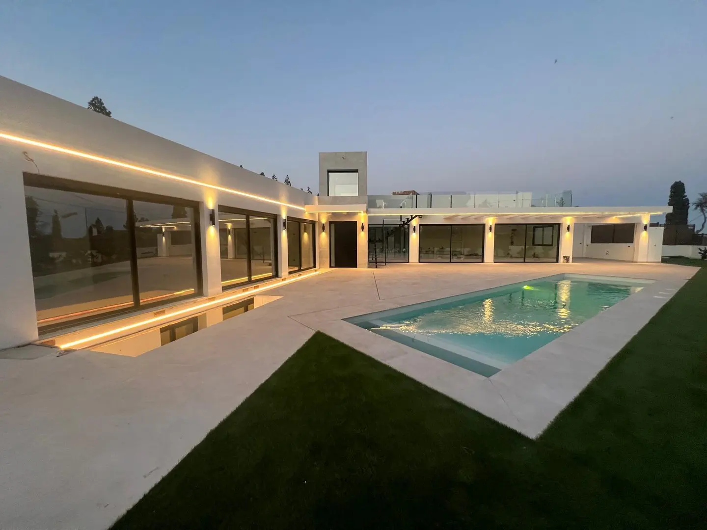 Modern white house exterior with a rectangular pool, large windows, and warm lighting at dusk.