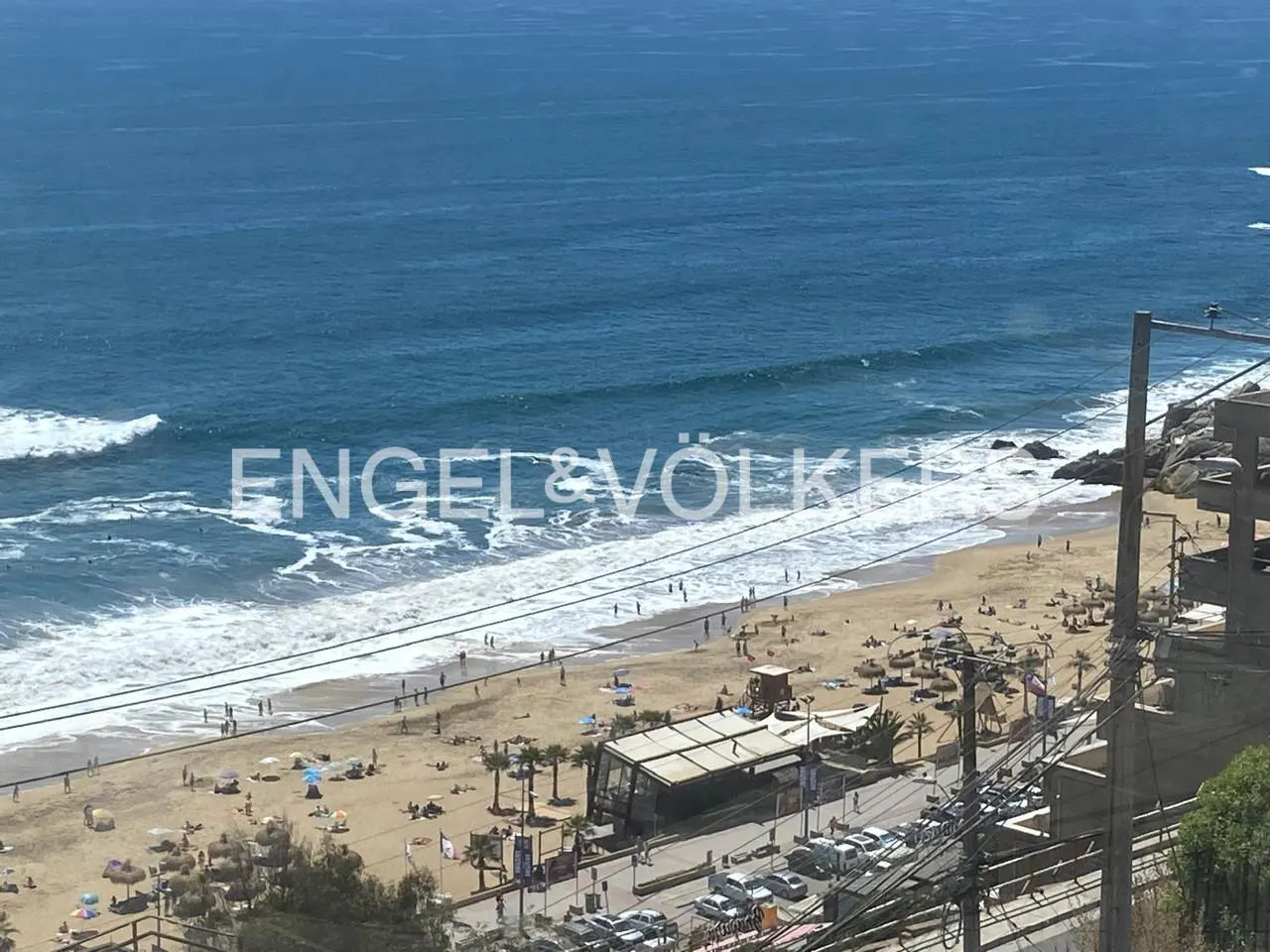 Ocean view of a crowded beach with blue water, white waves, and a sandy shore. Palm trees, buildings, and parked cars are visible.