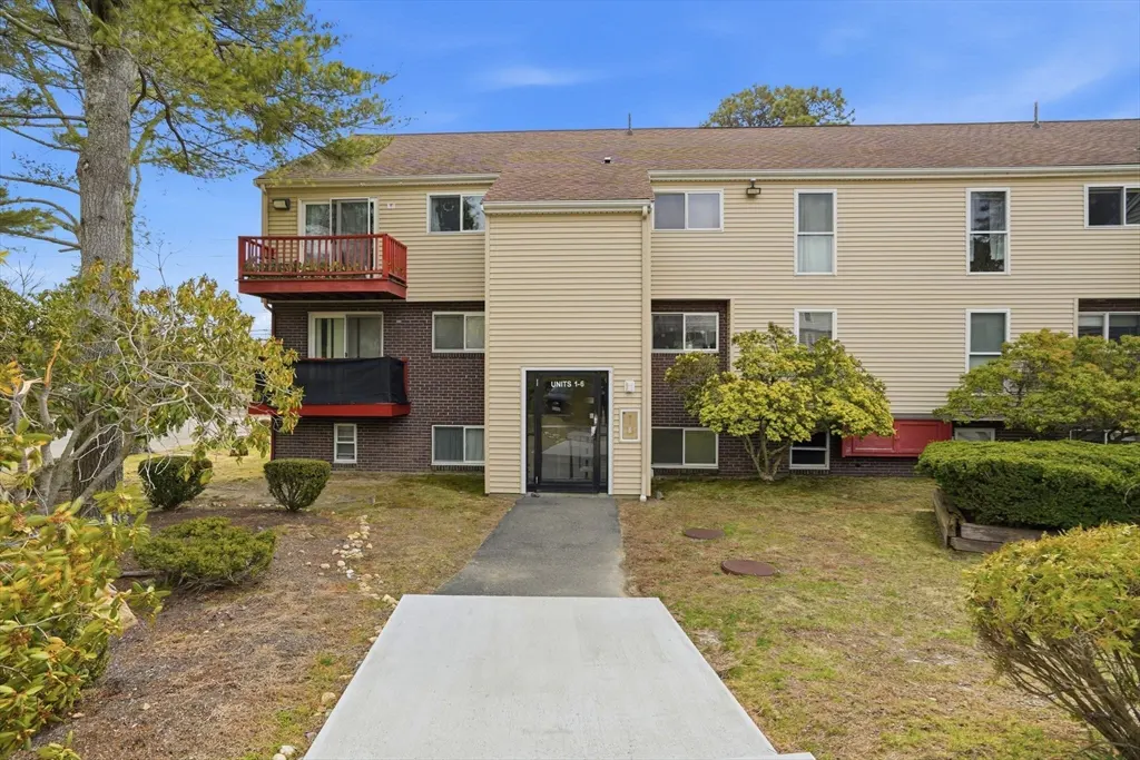 Exterior view of a two-story apartment building with beige siding and red balconies. A concrete walkway leads to the entrance door.