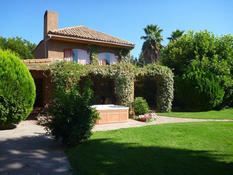 Exterior view of a brown stucco house with a tile roof, ivy-covered pergola, and a wooden hot tub on a green lawn.