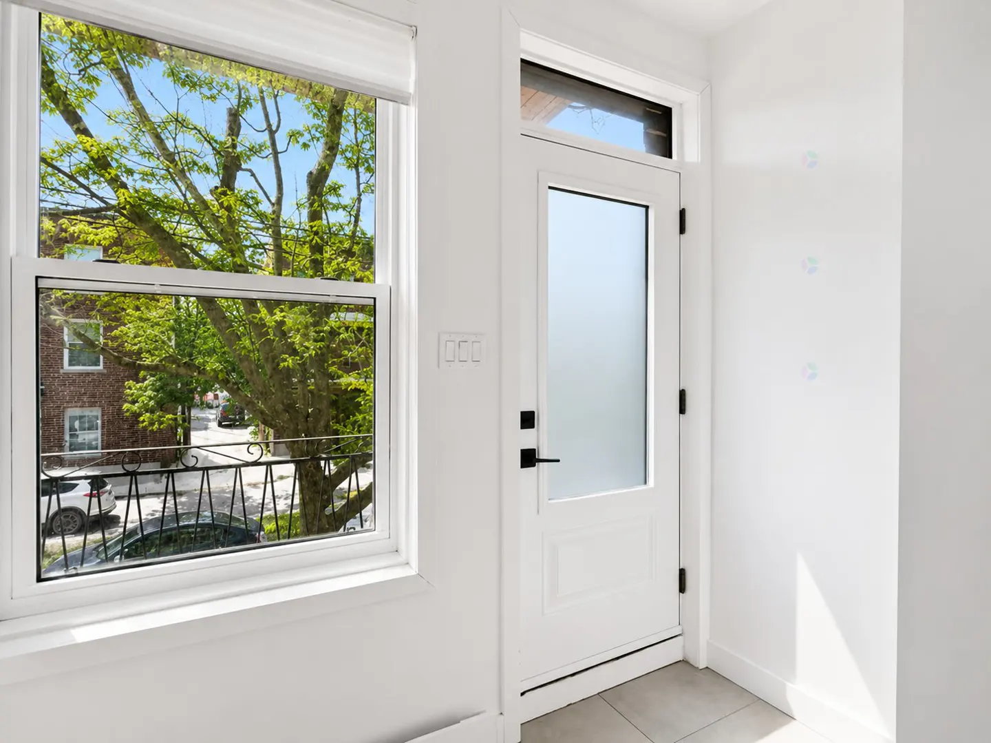 A bright room with a white door featuring a frosted glass panel next to a window overlooking a tree-lined street.
