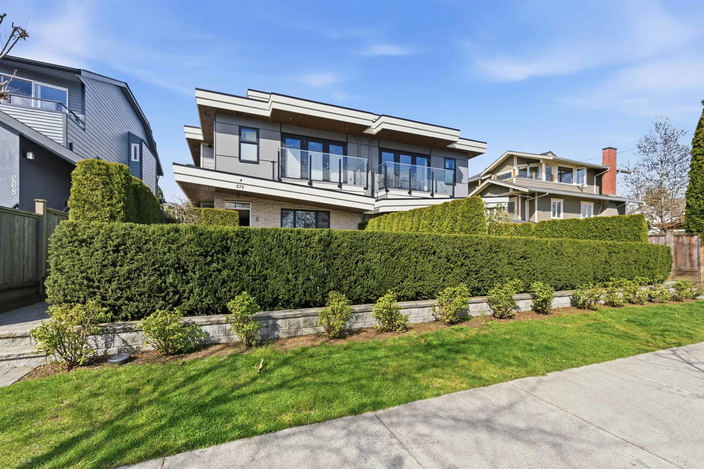 Modern two-story gray house with glass balcony railings, surrounded by a tall green hedge and a green lawn under a blue sky.