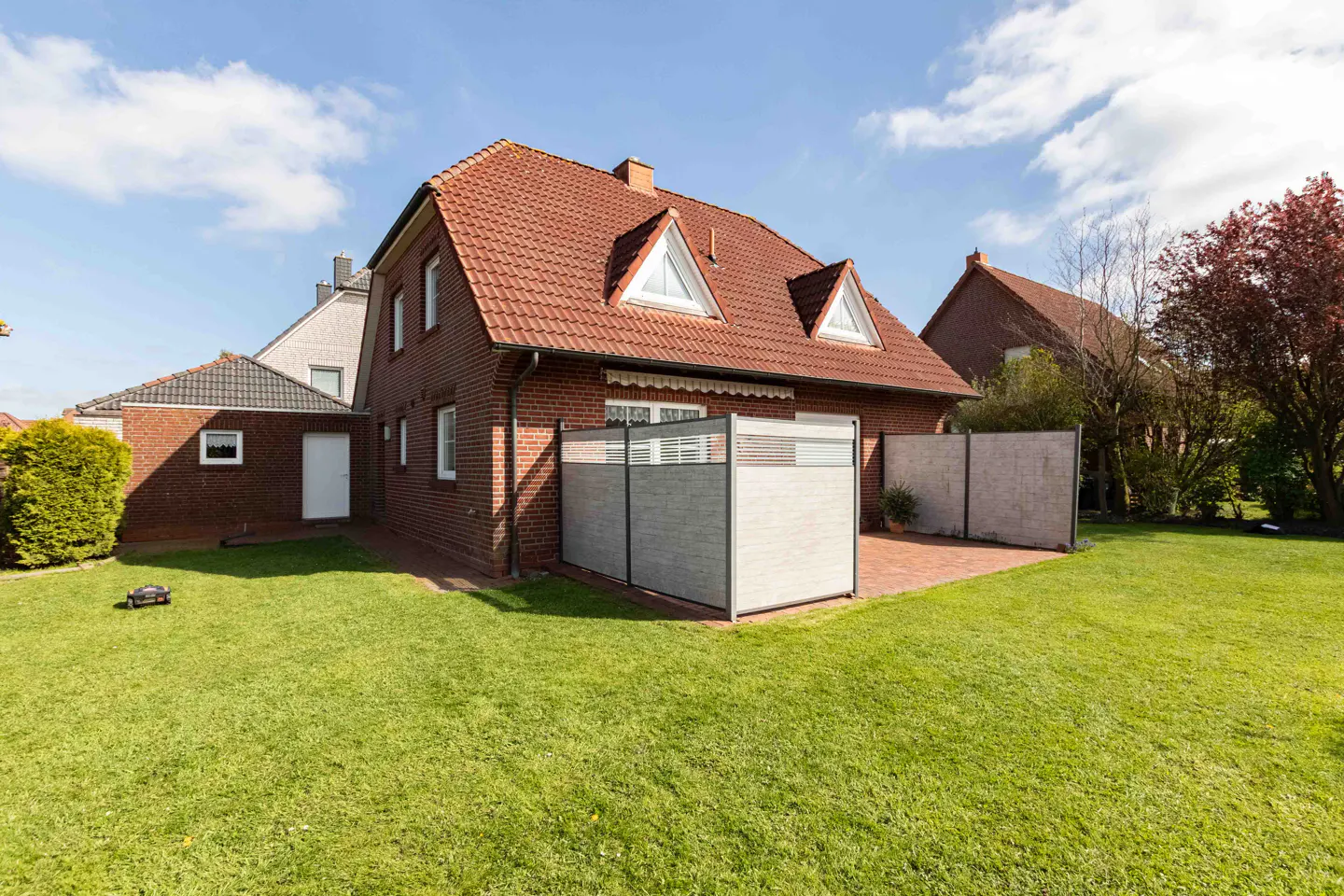 Backyard view of a two-story brick house with a red tile roof, green lawn, and privacy screens on a sunny day.