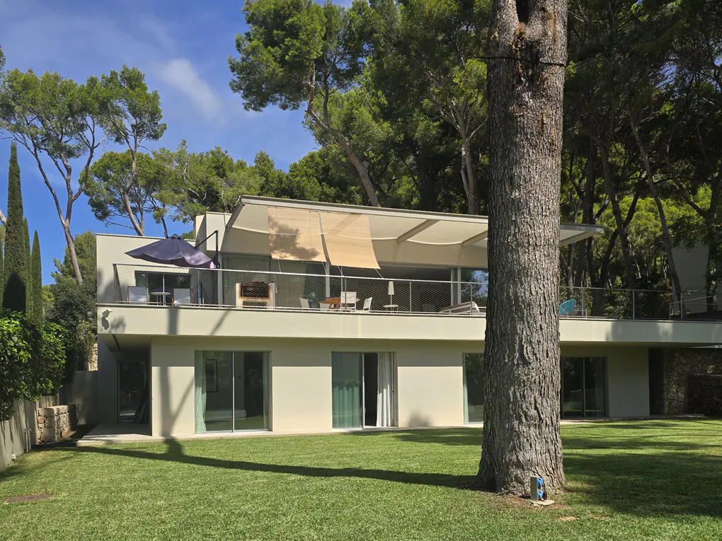 Modern two-story villa with a large balcony, outdoor seating, and a spacious green lawn surrounded by tall pine trees.