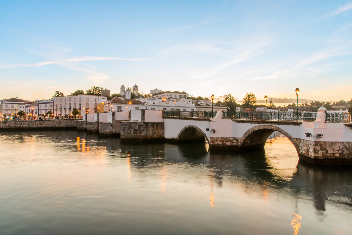 Bridge over calm river at dusk. White buildings line the shore under a blue sky. Street lamps illuminate the scene.