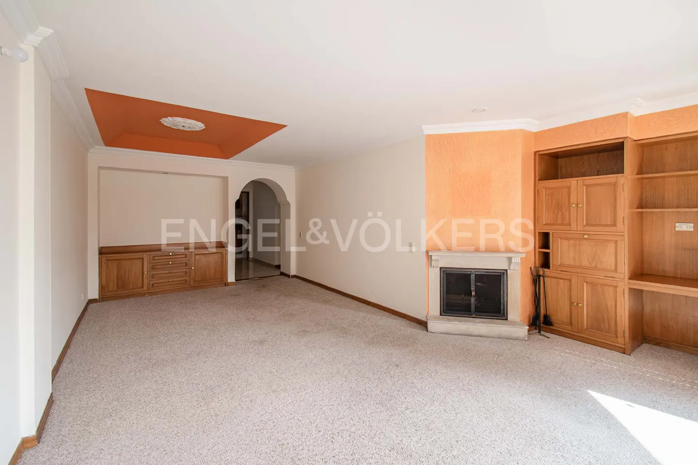 Spacious living room with beige carpet, built-in wooden cabinets, and a fireplace. Orange ceiling accent and archway visible.