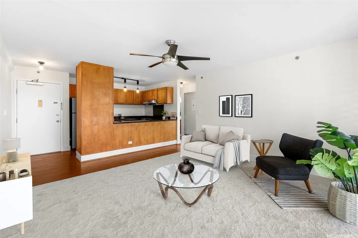 Open-concept living room with a white sofa, black armchair, glass coffee table, and wood-paneled kitchen in the background.
