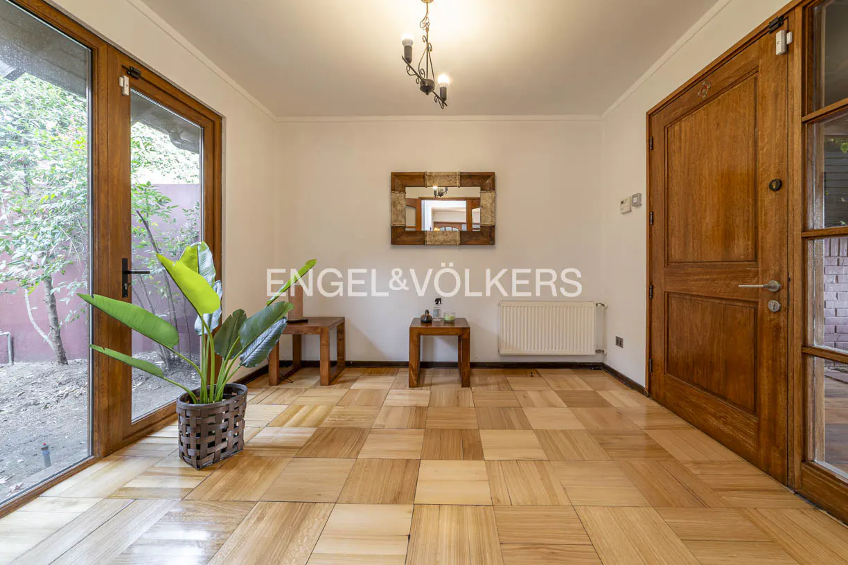 Entryway with wood-paneled floors, white walls, and a wooden door. A plant sits near a glass door to the left. A mirror hangs above two small tables.