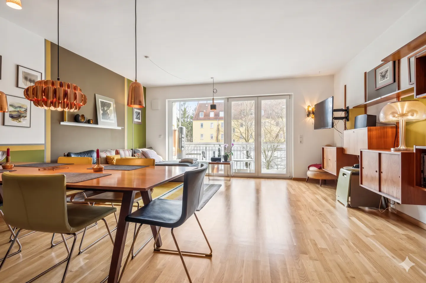 Bright, modern living and dining area with wood floors, table, chairs, and balcony doors. Copper pendant lights hang above the table.