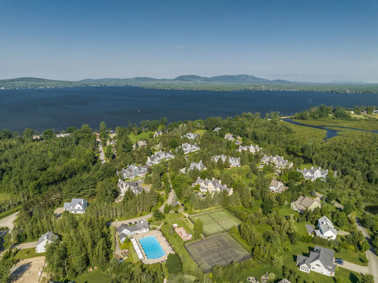 Aerial view of a lakeside community with houses, a pool, and tennis courts surrounded by green trees. Mountains are visible in the distance.