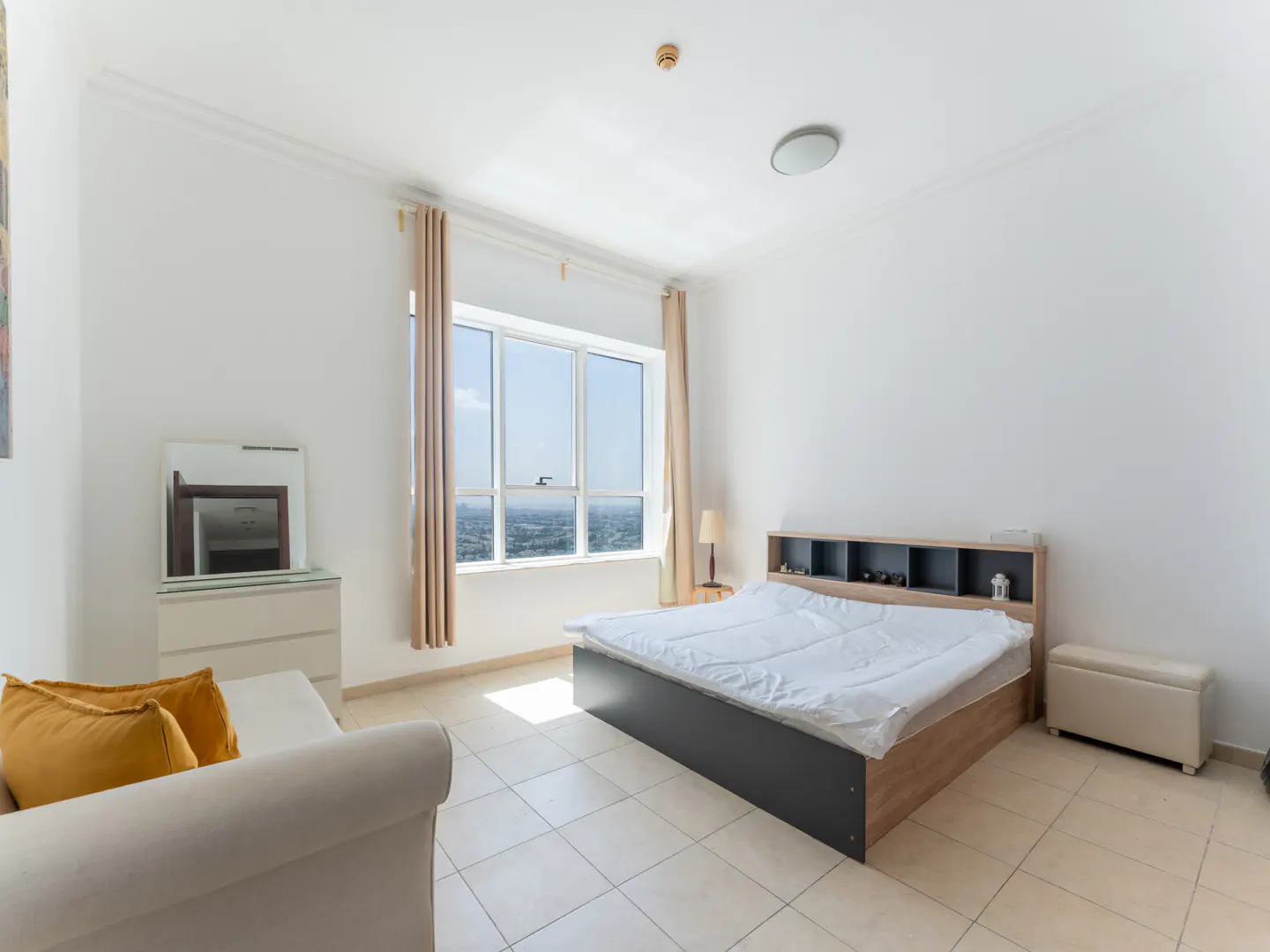 Bright bedroom with a large window showing a city view, a bed with a white comforter, and a beige sofa with yellow pillows.