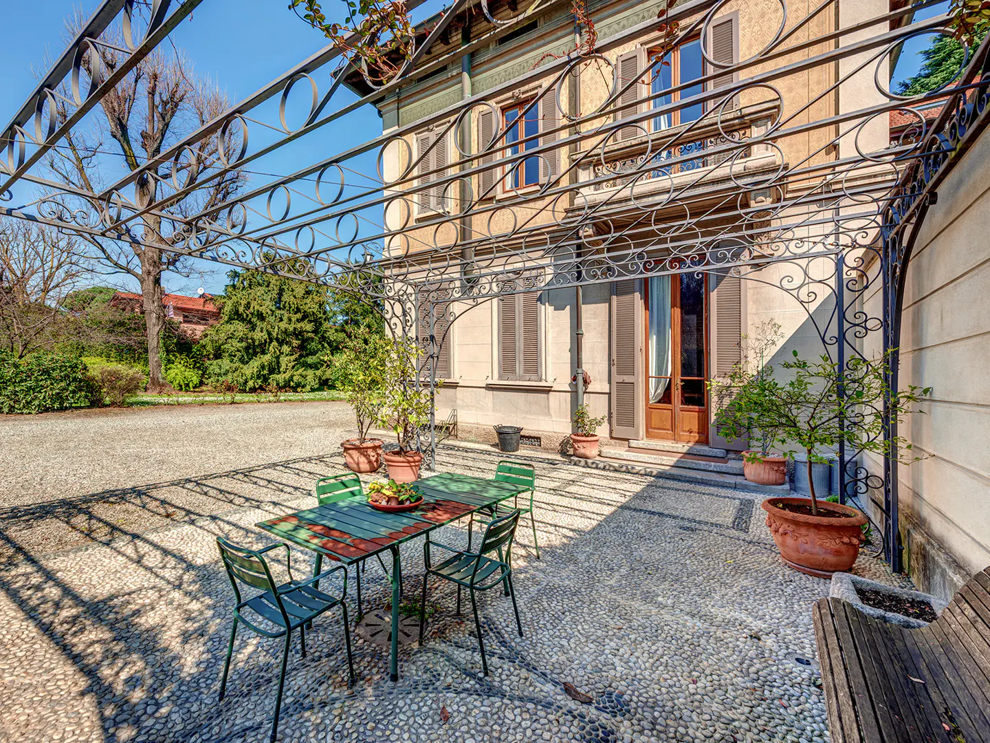 Outdoor patio with green table and chairs under a metal pergola. A stone path leads to a beige house with brown shutters.
