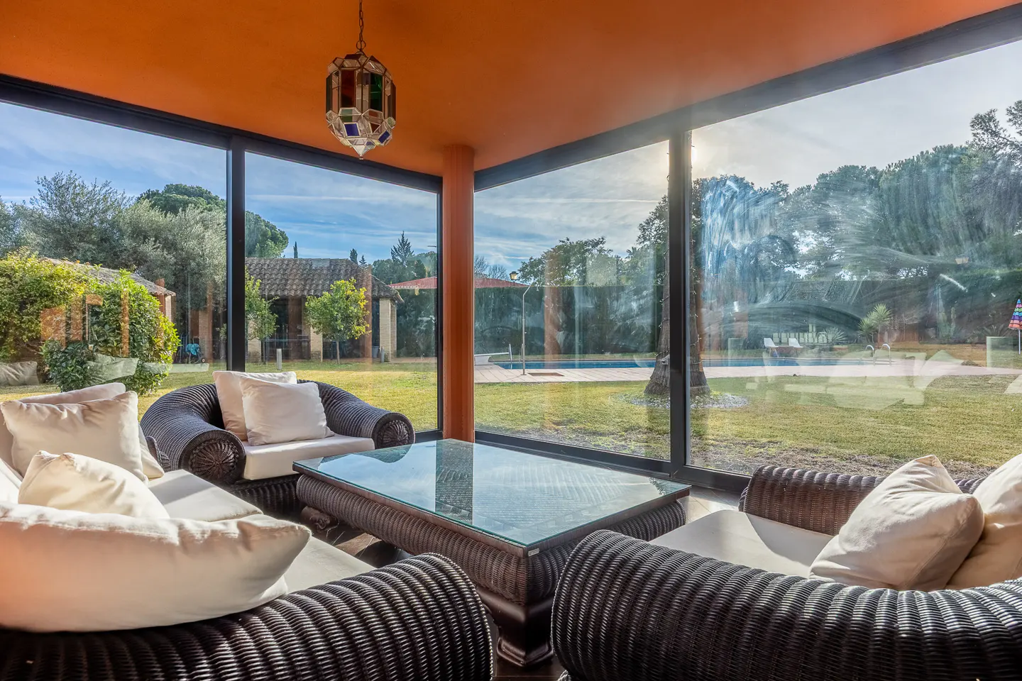 Sunroom with wicker furniture, white cushions, and glass table. Large windows overlook a green lawn, pool, and trees. Orange ceiling and colorful hanging lamp.
