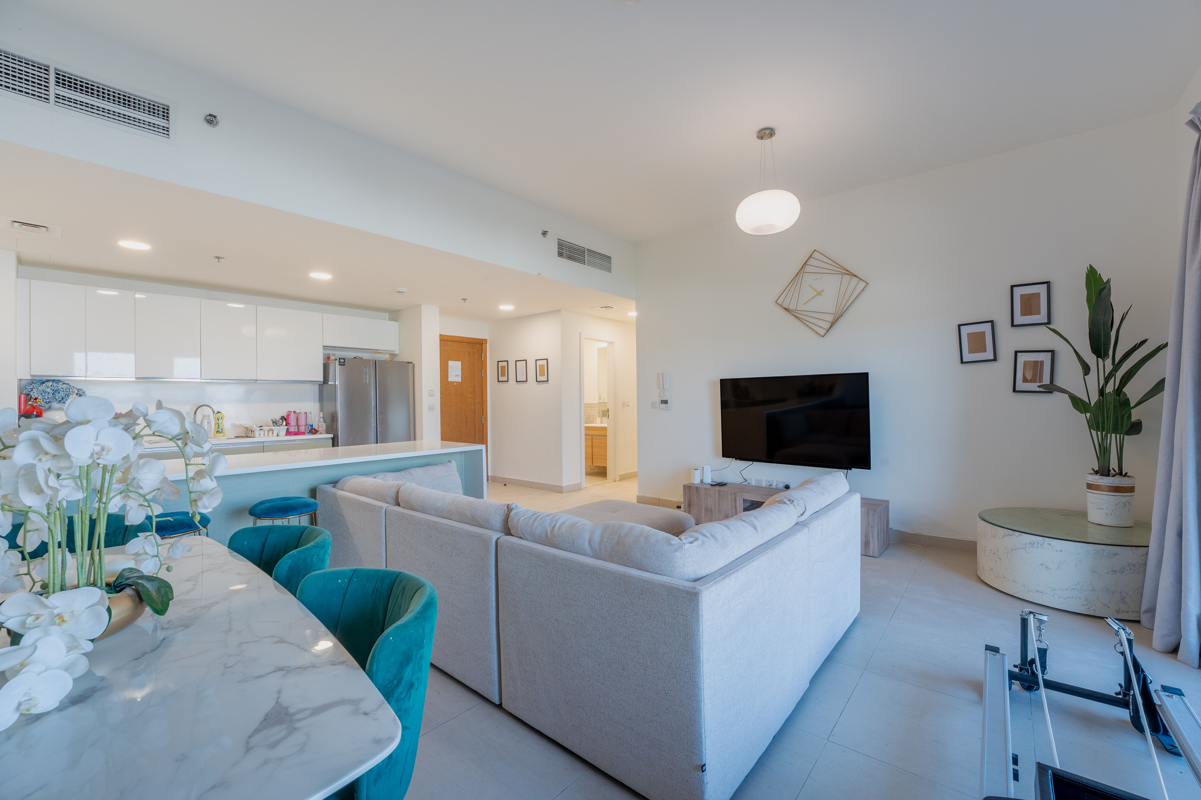 Bright, modern apartment interior with white walls, a large sectional sofa, and a marble dining table with teal chairs. A TV and plant add to the living space.
