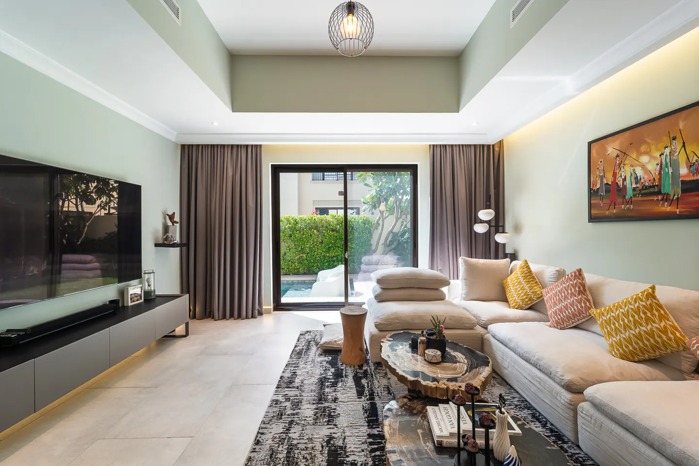 Living room with a large TV, beige sectional sofa with patterned pillows, and a coffee table with books. Sliding glass doors lead to a pool.