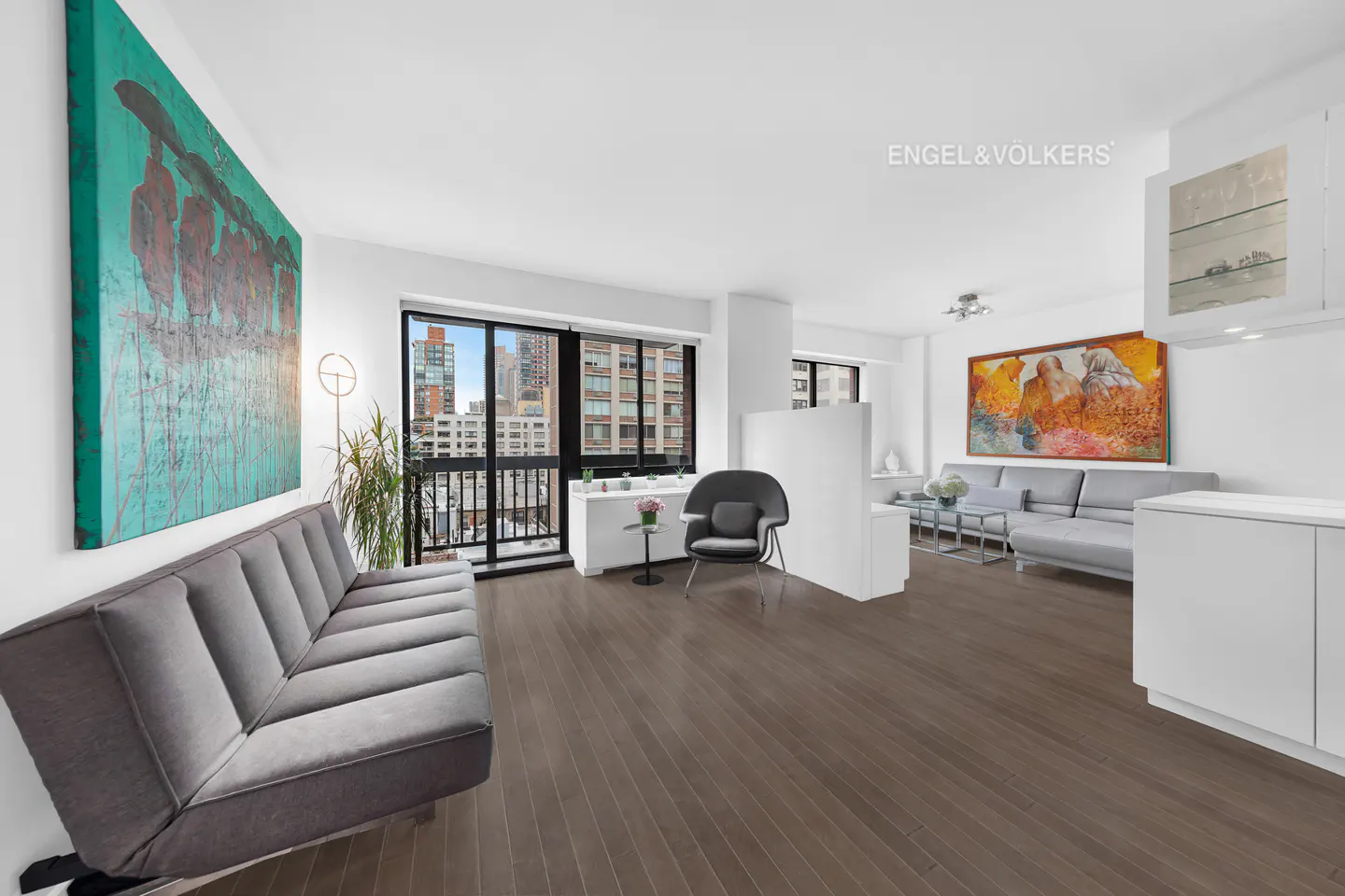 Bright, modern apartment interior with hardwood floors, gray sofas, and large abstract art. A city view is visible through the window.