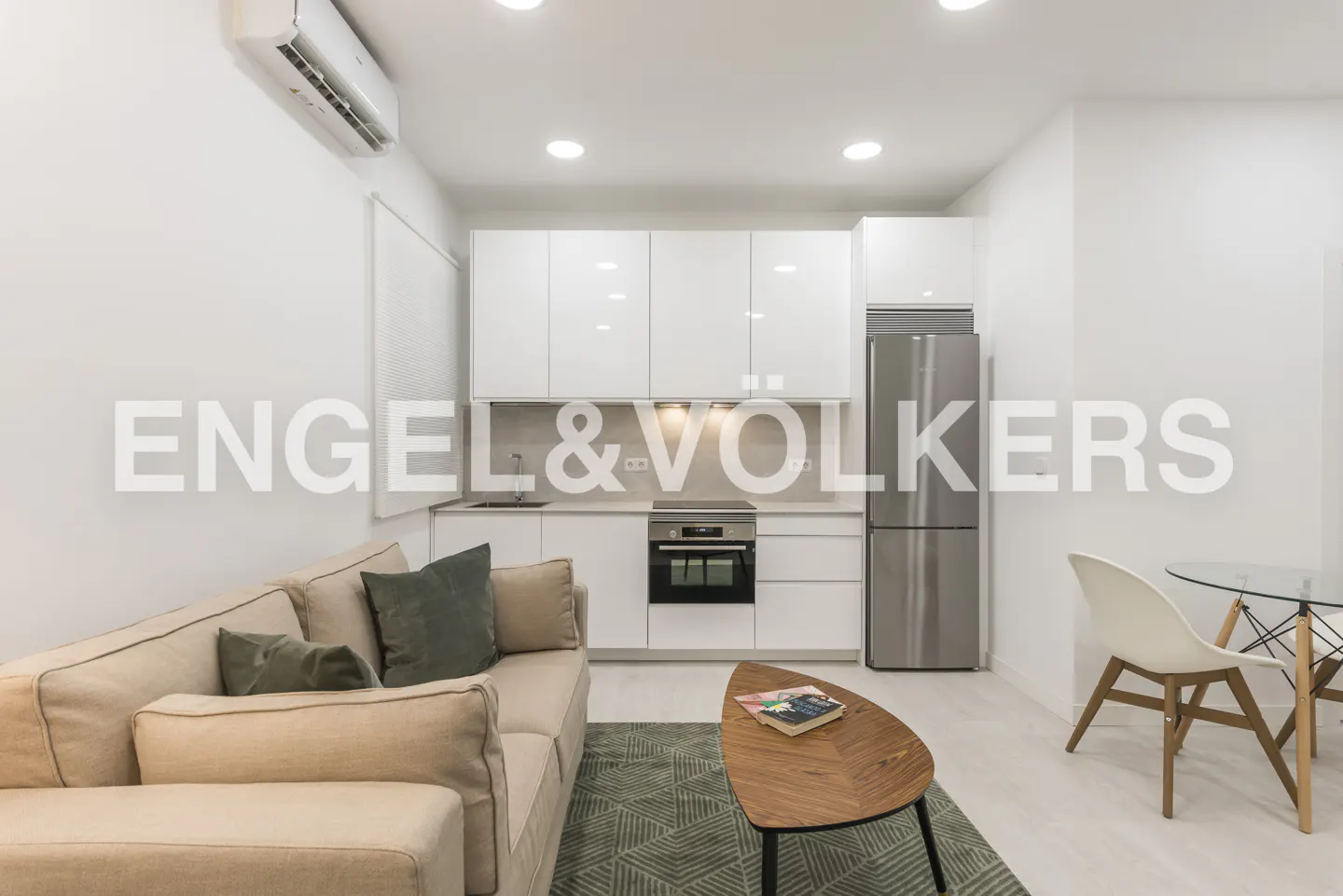 Bright, modern apartment interior with a beige sofa, wood coffee table, and white kitchen cabinets. Stainless steel refrigerator. Engel & Völkers logo overlay.