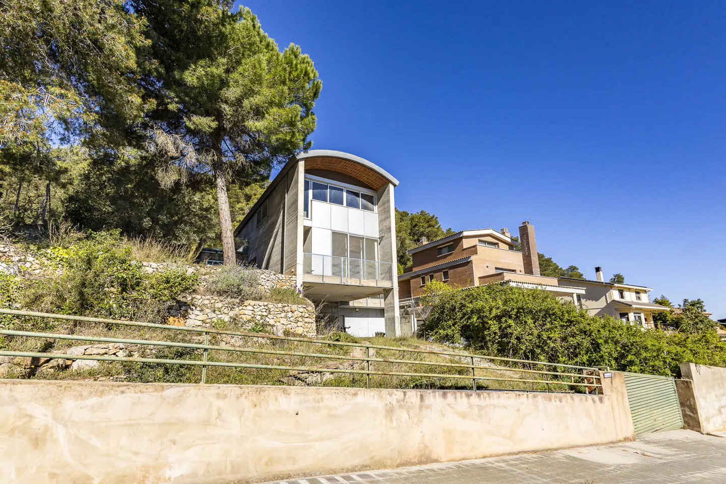Modern two-story house with a curved roof, set on a hillside with trees and other houses in the background.