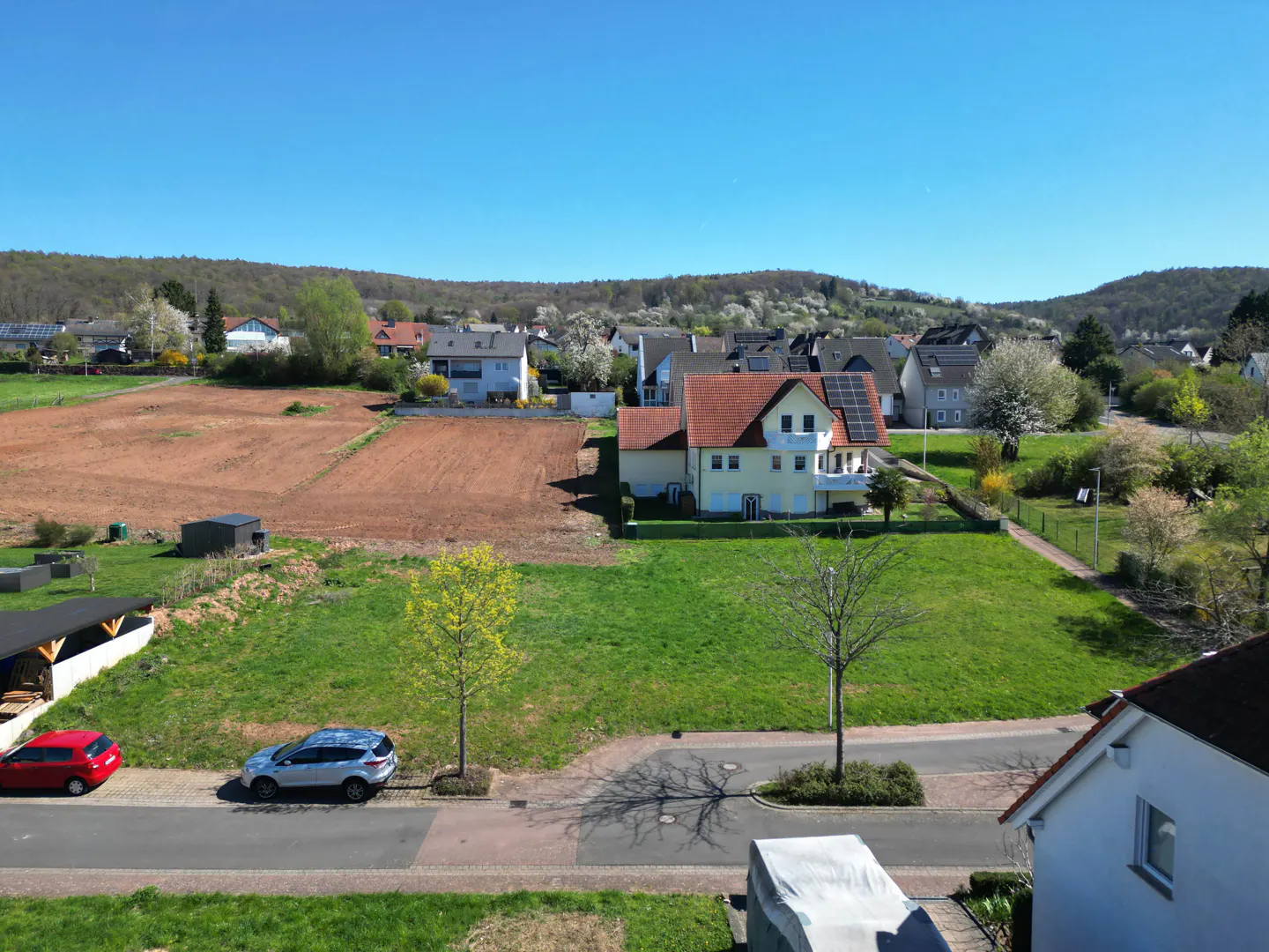 Aerial view of a yellow house with a red tile roof, solar panels, and a green lawn in a rural area on a sunny day.