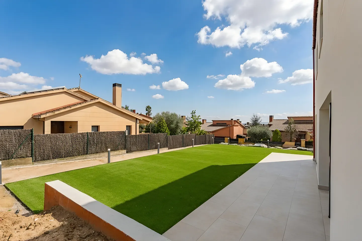 A backyard with green grass, a white tiled patio, and a view of houses under a blue sky with white clouds.