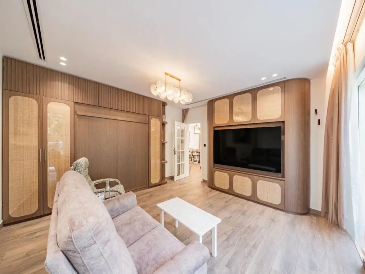 Living room with a gray sofa, white table, and a large TV set in a wooden cabinet. A glass door leads to another room.