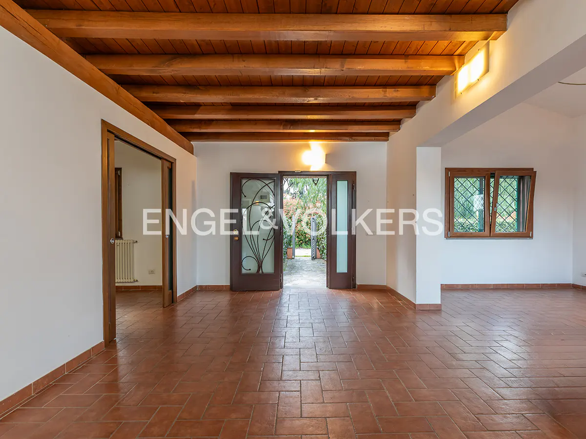 An interior view of a spacious room with terracotta tile flooring, white walls, and a wooden beamed ceiling. A doorway leads to a garden.
