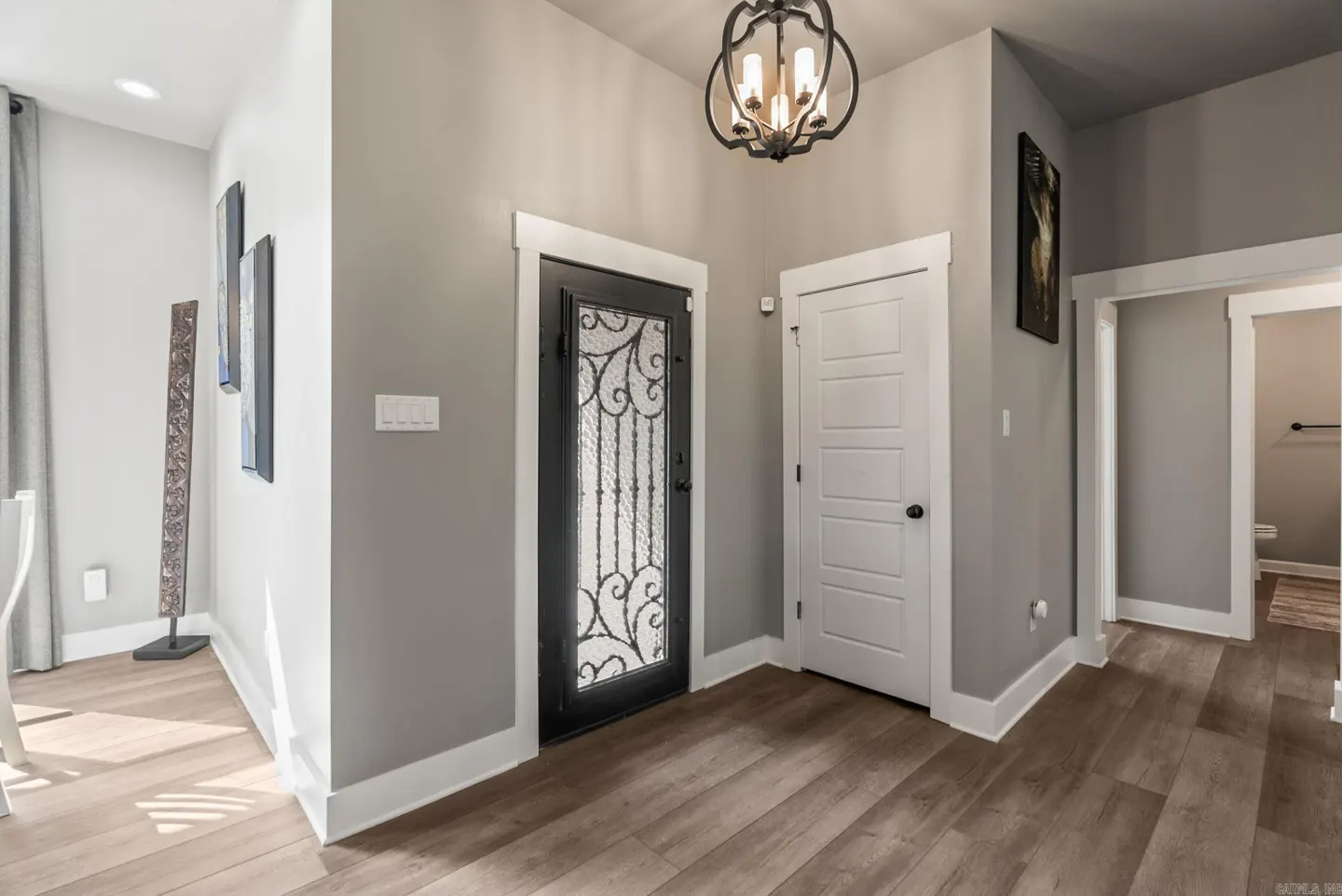 A gray foyer with wood floors, a black ornate front door, and a white interior door. A black chandelier hangs from the ceiling.