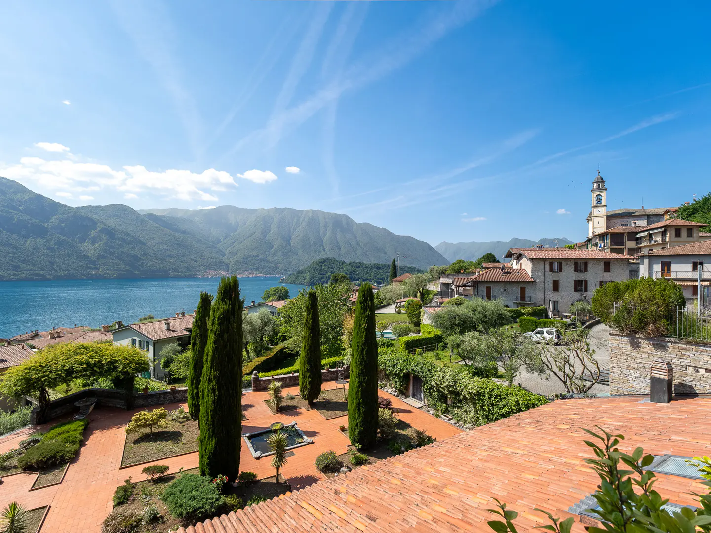 View of a terracotta-tiled terrace overlooking a lush garden with cypress trees, Lake Como, and a village with a bell tower.